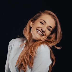 About Us Portrait of a cheerful woman with long blonde hair smiling against a black background.