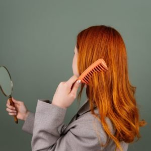 About Us Red-haired woman using a comb while holding a mirror, focusing on hair care and beauty.