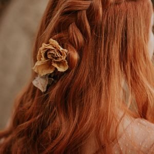 About Us Red-haired woman with a braided hairstyle adorned with a dried flower, captured in a soft, warm tone.