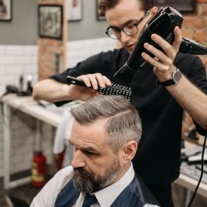 About Us Barber styling a bearded client's hair using a blow dryer and brush in a trendy salon.